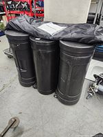 Three closed black cylindrical wheeled hard plastic cases with a folded fabric bag on top