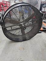 Full view of large black industrial floor fan with metal blades and mesh guard, showing missing wheels and condition