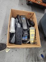 Top view of a cardboard box filled with a variety of remote controls in mixed colors and conditions, showing shape and button details for many remotes.