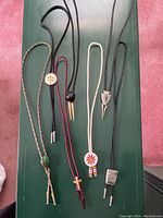 Photo showing seven vintage bolo ties arranged side by side on a green surface, highlighting variety in colors and clasp designs.