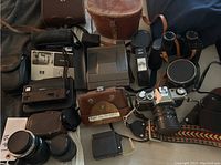 Various vintage cameras and accessories spread out on a surface, showing leather cases, different camera types, lenses, and accessories.