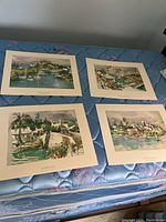 Four Cecile Johnson Bermuda prints laid out on a blue floral mattress, showing different scenic views with boats, bridges, and buildings.