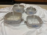 Four Corningware casserole dishes on white cloth, showing Cornflower Blue and Provincial Blue designs with glass lids.