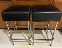 Front view of two black cushioned square barstools with metal bases and footrests, positioned side by side on a floor.