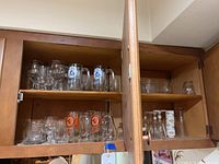 Four cabinet shelves with various glassware, featuring beer mugs with Senators and Baltimore Orioles logos, stemware, and tumblers.