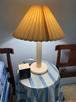Photo of a vintage cream pleated lamp with a wide base on a small table, next to a black Sony Dream Machine clock and another small device.