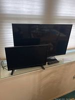 Samsung 32-inch TV behind Insignia monitor on glass table near window with beige shade.