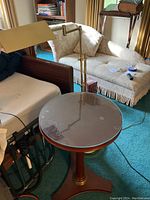 Full view showing brass standing lamp attached to the side of the round table with glass top on triangular wooden base with brass accents