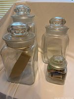 Four vintage glass storage jars of varying sizes with decorative round lids. The smallest jar contains vintage matchboxes. All jars are displayed on a flat surface with neutral background.