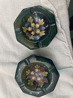 Top view of two octagonal William Moorcroft ashtrays showing detailed Clematis floral designs in yellow, blue, and purple on green glaze.