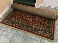 Full view of the Tommy Bahama Estilo wool rug partially rolled on a tiled floor showing red central field with floral and palm tree motifs and beige fringed edge.