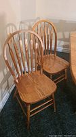 Two oak chairs shown side-by-side with contoured seats and vertical spindle backs in natural finish on carpeted floor.