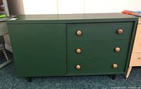Front view showing dark green dresser with three wooden pull drawers and a cabinet door on the left