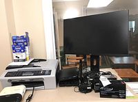 Overview of Brother MFC-240C multifunction printer with spare ink cartridges, Sony Dream Machine clock radio, router, Enerwatt battery box, and Monoprice monitor with cords on tabletop.