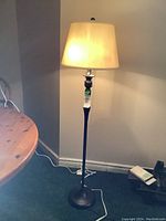Floor lamp with dark metal base and cream fabric lampshade, lit and standing next to a wooden table on carpeted floor.