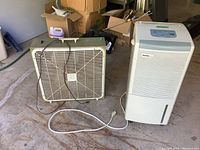 Both the Danby air humidifier and the Rotoe electric box fan shown together on a garage floor with some boxes in the background.