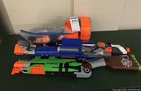Three Nerf blasters laid out on a table with a resealable bag of orange darts and two Nerf magazines.