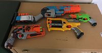Four Nerf toy guns arranged on a brown surface showing various colors and accessories with some foam darts.