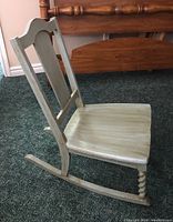 Side view of wooden rocking chair showing ornate spiral turned front leg and carved backrest with distressed cream finish.