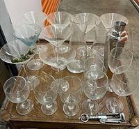 Top-down view of assorted clear glass barware including martini glasses, wine glasses, a cocktail shaker, and metal bottle openers.