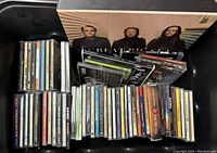 Photo showing a variety of music CDs stacked and spread, including a visible Greatest Hits compilation and other assorted artist albums in jewel cases and cardboard sleeves.