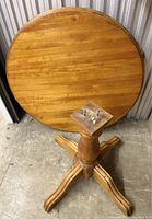 Photo of a round wooden dining table top detached from its pedestal base, standing on a concrete floor in a storage unit.