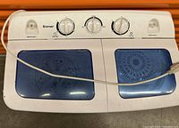 Top view showing control panel with three dials and separate blue lids for washer and spin dryer compartments. Includes water inlets and power cord.