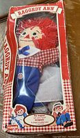 Photo of Raggedy Ann doll inside original Playskool box with visible red yarn hair, red and white check shirt, and blue pants.