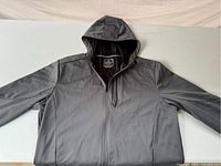 Full front view of the grey men's jacket with sleeves outstretched, hood up, showing zipper and chest pocket.