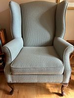 Front view of the armchair showing light textured fabric, wingback design, and wooden curved front legs.