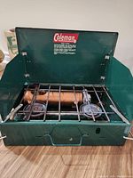 Front view of the Coleman green two-burner camping stove with hinged lid open showing burners, metal grate, and manual fuel pump.