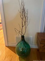 Green glass vase with a rounded bulbous base and narrow neck, sitting on the floor with natural twigs inside.
