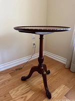 Round wooden accent table with carved scalloped edge top, turned pedestal, and carved claw feet on hardwood floor near wall outlet.