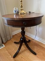 Side angle of the round wooden table showing its pedestal and four claw-foot legs along with single drawer pull.