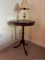 Wooden round table with a polished dark wood finish, a central turned pedestal leg, and three outward curved legs with brass tips, photographed against a cream wall with a lamp on top (lamp not included).