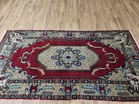 Full view of rectangular oriental style rug on wooden floor showing central medallion and border pattern in red, beige, blue and black colors