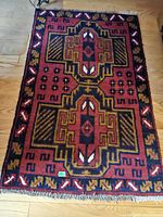 Top-down photo of a rectangular decorative rug with a geometric central pattern and fringe edges on a wooden floor.