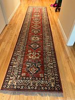 Photo of a long runner rug placed in a hallway with a light wood floor. The rug has a deep red field with multiple diamond and floral motifs, and intricate borders in cream, navy, and other colors.