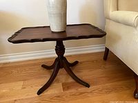 Photo of a dark wood rectangular low side table with scalloped edges and a turned pedestal base with four curved legs. A ceramic vase is placed on the table but not included in the lot.