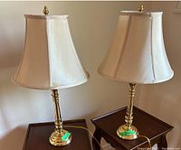 Pair of brass table lamps with cream colored fabric lampshades shown on wooden furniture.