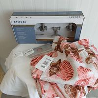 VCNY Home Shell Treasure shower curtain folded showing coastal shell pattern in rust, brown, pink on light background with Moen Metry BH5033BN 3-piece bath accessories kit box and accessories on table.