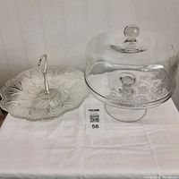 Photo showing the clear glass etched pedestal cake stand with dome lid and the Indiana Clear Glass Pebble Leaf one-tier tray side by side on a white cloth.