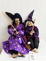 Two witch figurines sitting next to each other, dressed in vibrant purple and black robes, each holding a broomstick.