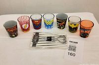 Wide shot showing seven shot glasses with various Marvel and DC Comics characters and logos on a white surface.