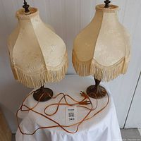 Pair of Victorian lamps on a table, showing ivory cream floral-patterned shades with long matching fringe, brass bases, and attached power cords.