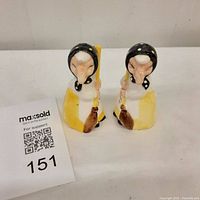 Front view of the pair of witch salt and pepper shakers, showing detailed faces and full bodies holding brooms with a MaxSold lot card beside them.