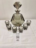 Full set showing decanter with stopper and six faceted glasses arranged in front, all made of smokey glass with geometric design.