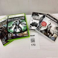 Four video game cases grouped, two Xbox 360 games on left (Supreme Commander, Bioshock) and two PS3 games on right (Battlefield Bad Company 2, Assassin's Creed III).