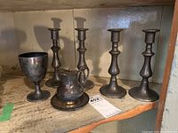 Wide shot showing all items in the set: 4 pewter-style candlesticks, goblet, creamer with tray, on wooden shelf.