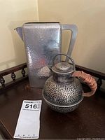 View of the dimpled tin rectangular pitcher and the round teapot with wicker handle on wooden surface.